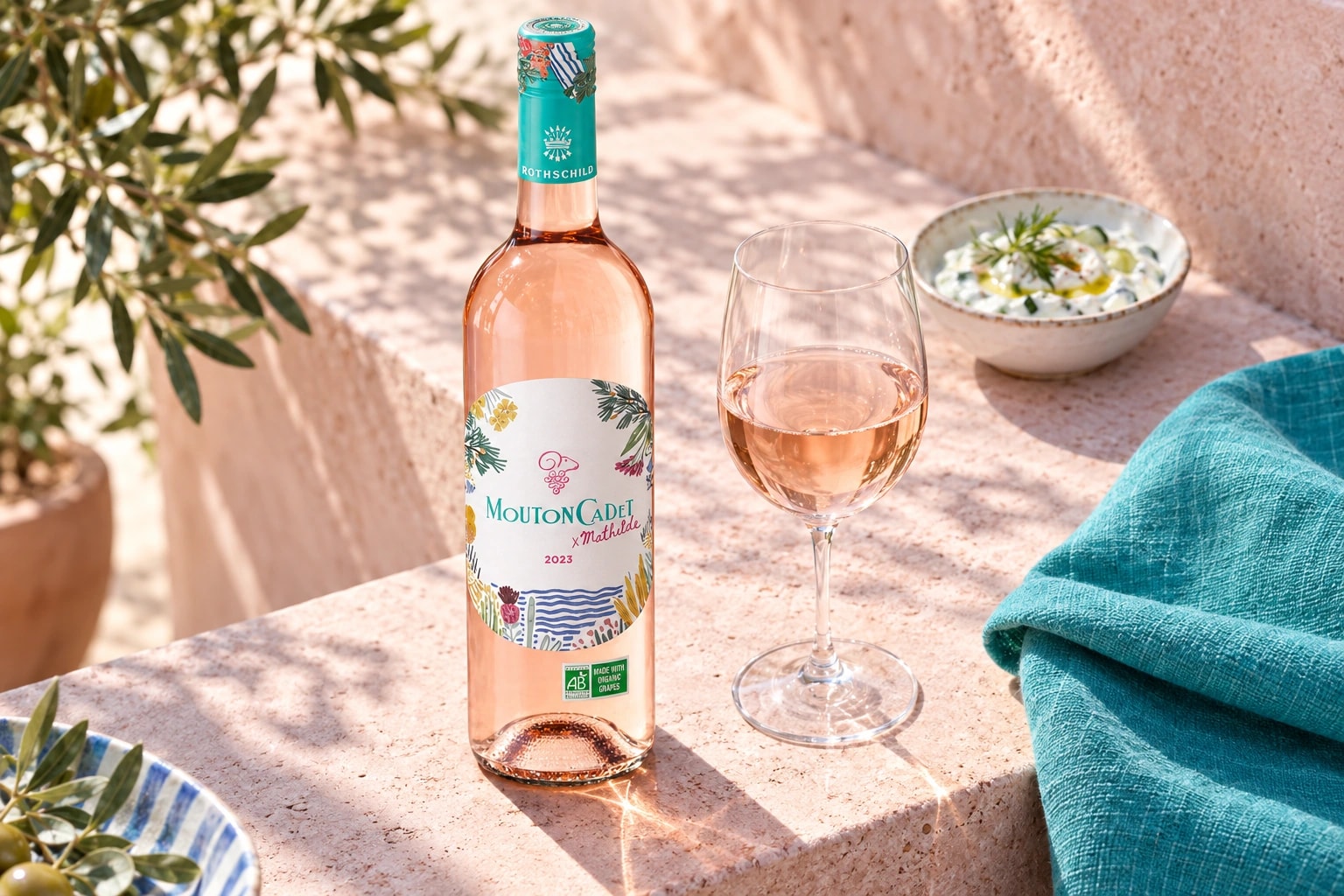 Mouton Cadet Rosé x Mathilde on pink stone beside a rosé glass and turquoise linen