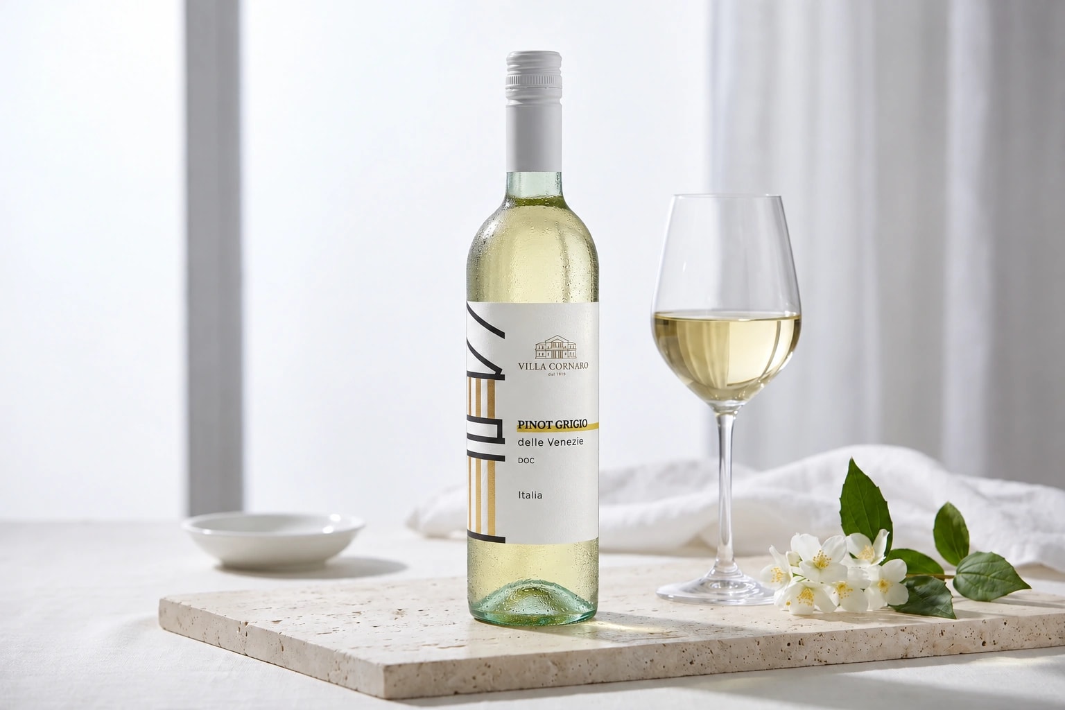 Villa Cornaro Pinot Grigio on pale travertine beside a glass of straw-yellow wine and a sprig of jasmine